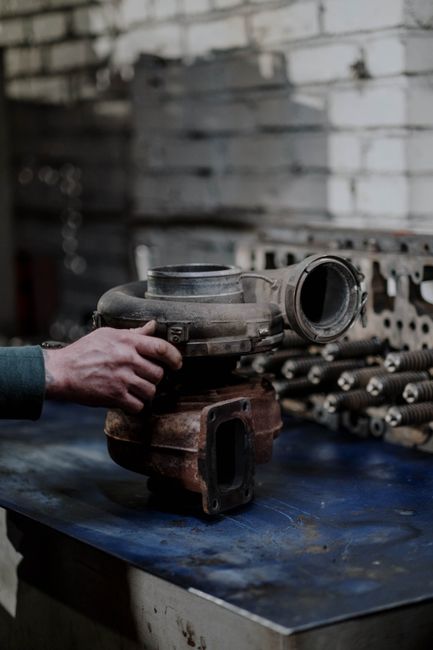 A person's hand is gripping a large, metallic turbocharger component, which is placed on a dark blue workbench inside a workshop or storage area. In the background, there are rows of metal bolts and t