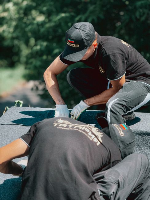 Two individuals wearing black uniforms and caps with the logo of Tomsmanandvan.co.uk are engaged in a home relocation or furniture transport task outdoors on a paved surface surrounded by greenery. On